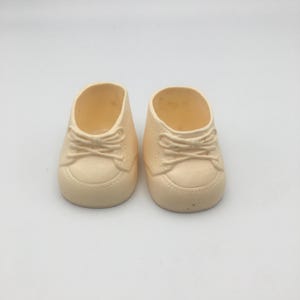 Könnte beinhalten: Ein Paar kleine, cremefarbene Puppenschuhe mit einem geformten Schnürdesign, einer abgerundeten Spitze und einer glatten Oberfläche. Diese Miniaturschuhe haben eine einfache, solide Konstruktion mit sichtbaren Details, die herkömmliches Schuhwerk imitieren.