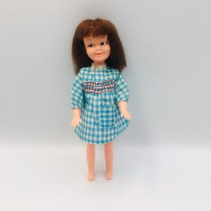May include: A doll with shoulder-length brown hair and painted facial features, wearing a light blue and white gingham dress. The dress features a smocked bodice with red stitching, short sleeves, a small front pocket, and a white lace collar.