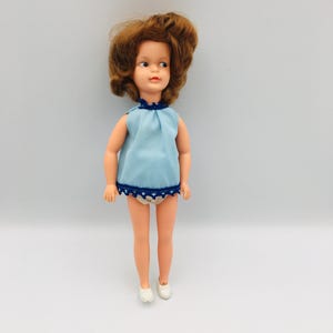 May include: A doll with shoulder-length auburn hair, blue eyes, and a light skin tone. The doll wears a sleeveless light blue top with a dark blue scalloped trim at the neckline and hem, paired with white undergarments and simple white shoes.