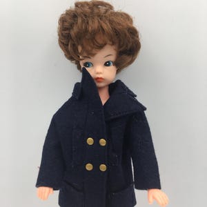 Puede incluir: Una muñeca vintage con cabello castaño y ojos azules que lleva un abrigo cruzado azul marino con botones dorados. El abrigo de la muñeca tiene un diseño clásico con cuello con solapas y bolsillos.