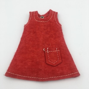 May include: A small, red felt dress with white stitching details. The dress features a small pocket and white stitching around the neckline and hem. The dress is sleeveless and has a simple, A-line shape.