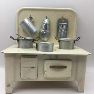 以下が含まれることがあります： クリーム色のヴィンテージおもちゃのキッチンセット。金属製の鍋、おろし金、計量カップが含まれています。ストーブには2つのバーナーと引き出しがあります。セットにはトウモロコシ型の型が含まれています。