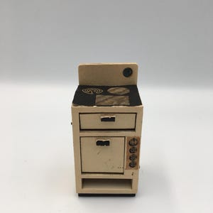 Puede incluir: Una estufa de juguete en miniatura de color crema con una encimera y un horno negros. La estufa tiene dos cajones y una pequeña balda en la parte inferior. La encimera presenta dos quemadores y un diseño decorativo. Un pequeño emblema redondo está en la parte superior.