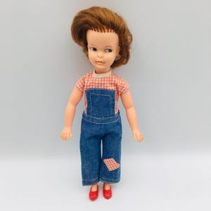 Könnte beinhalten: Eine Puppe mit kurzem rotbraunem Haar und blauen Augen, bekleidet mit einem rot-weiß karierten Kurzarmshirt unter einer blauen Jeanslatzhose. Die Latzhose hat einen kleinen rot-weiß karierten Flicken am linken Bein. Die Puppe trägt rote Schuhe.