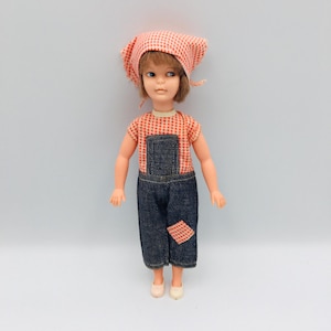 May include: A plastic doll with brown hair wearing a red and white checkered headscarf, a matching short-sleeved shirt, and dark blue denim overalls. The overalls feature a small red and white checkered patch on the left leg. The doll wears light-colored shoes.