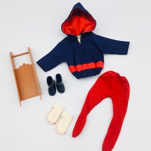 May include: A vintage doll winter outfit set. The set includes a navy blue hooded jacket with a red lining and trim, red pants, black boots, white mittens, and a brown wooden sled. The outfit is designed for a doll.