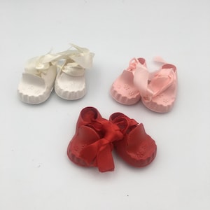 Puede incluir: Tres pares de zapatos para muñeca en blanco, rosa claro y rojo. Cada par presenta una punta redondeada con un patrón floral moldeado, un borde festoneado a lo largo de la suela y cordones de satén a juego atados en la parte superior.