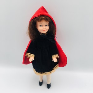 May include: A vintage doll in a black velvet dress with gold trim and a red hooded cape. The doll has brown hair, white tights and black shoes. The doll is set against a white background.
