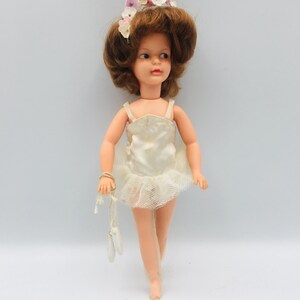 May include: A doll with brown hair styled in a bouffant, wearing a floral headpiece and a white satin leotard with a tulle skirt. The doll holds a pair of white ballet slippers by a string in one hand and has painted facial features.