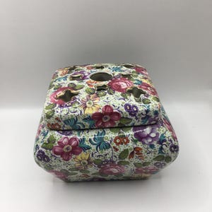 May include: A floral-patterned ceramic container with a lid. The container is decorated with a variety of colorful flowers, including pink, purple, and yellow blooms, set against a light green background. The lid has several openings.