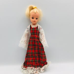 Könnte beinhalten: Eine Vintage-Puppe mit blonden Haaren im Pferdeschwanz. Die Puppe trägt ein rot kariertes Kleid mit weißer Spitzenborte und einem weißen Spitzentop. Das Kleid hat einen Rüschensaum. Die Puppe ist ein Sammlerstück.