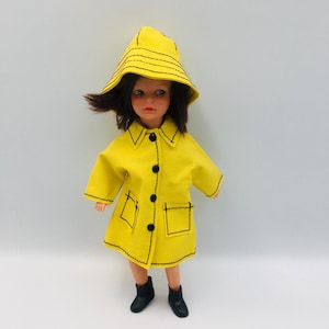 May include: A vintage doll dressed in a bright yellow raincoat and matching hat with black stitching. The doll has dark hair and wears black boots. The coat features black buttons and pockets. The doll is set against a white backdrop.