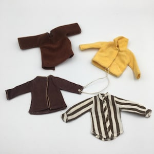 Puede incluir: Una colección de cuatro camisas en miniatura para muñecos que incluye un jersey marrón liso, una chaqueta amarilla con capucha, una camisa abotonada de color marrón oscuro con costuras visibles y una camisa de manga larga con rayas verticales.