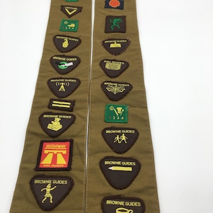 Peut inclure: Une écharpe beige avec deux colonnes d'écussons brodés. Les écussons présentent divers insignes de Brownie Guides et Girl Guides, tels qu'une ampoule, une toile d'araignée, un ours en peluche, un crayon, une tasse à thé et un panneau HIGHWAY GIRL GUIDES.