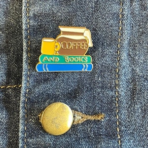 May include: Gold enamel pin featuring a brown coffee pot, a yellow mug, and three blue and green books. The text on the pin reads "Coffee and Books".
