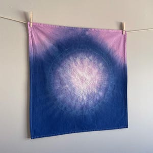 May include: A square, tie-dye textile in shades of blue, purple, and pink, suspended from a washing line with wooden pegs. The design features a central, lighter circular pattern, with darker hues radiating outwards.
