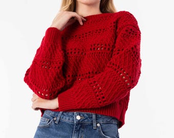 May include: A red knit sweater with a round neckline and long sleeves. The sweater has a textured pattern with openwork details. The model is wearing blue jeans. The background is white.