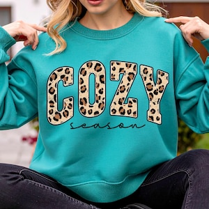 May include: Teal sweatshirt with the word "COZY" in leopard print letters and the word "season" in a cursive font. The sweatshirt has long sleeves and a crew neck.