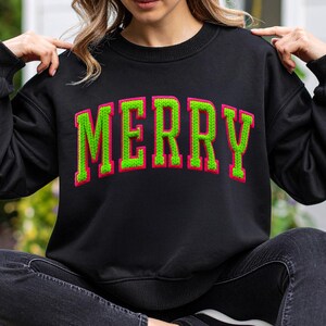May include: Black sweatshirt with the word "MERRY" in large, green, embroidered letters with a pink outline. The sweatshirt has a crew neck and long sleeves. The person is wearing black jeans and black sneakers.