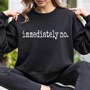 Puede incluir: Sudadera negra con la frase "immediately no." en blanco. La sudadera de cuello redondo y manga larga se usa con jeans negros y zapatillas negras. La persona está sentada con las piernas cruzadas.