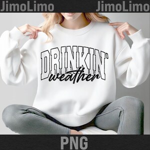 May include: White sweatshirt with the text "DRINKIN' weather" in a black, distressed font. The sweatshirt has a crew neck and long sleeves. The model is wearing grey jeans.
