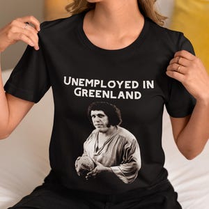 Könnte beinhalten: Schwarzes T-Shirt mit dem weißen Text "UNEMPLOYED IN GREENLAND" über einem Schwarz-Weiß-Bild eines Mannes, der einen Ball hält. Das T-Shirt hat kurze Ärmel und einen Rundhalsausschnitt.