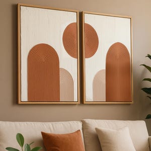 Könnte beinhalten: Zwei gerahmte abstrakte Kunstwerke mit strukturiertem weißem Hintergrund und terrakottafarbenen geometrischen Formen. Die Kunst hängt über einem beigen Sofa mit passenden Kissen und einem runden Couchtisch aus Holz mit einer Terrakotta-Vase.