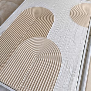 May include: Textured abstract art piece in a white frame. The artwork features a white background with beige, arched, textured shapes. The shapes have a ridged texture, creating a sense of depth and dimension. The piece is a modern, minimalist design.