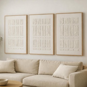 May include: Three framed wall art pieces with a textured, cream design. The artwork hangs above a beige sofa with matching cushions. A wooden side table with a woven lamp and a small bowl on a coffee table complete the living room setting.