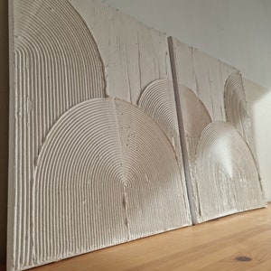 Op de afbeelding: Twee witte, textuurrijke wanddecoratiepanelen met een reliëfdesign van concentrische cirkels, die lijken op een reeks bogen.
