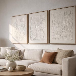 Peut inclure: Trois œuvres d'art abstraites encadrées avec des surfaces texturées blanc cassé sont accrochées au-dessus d'un canapé sectionnel beige. L'ensemble de salon comprend une table basse ronde en bois, des coussins décoratifs et une lampe de table.