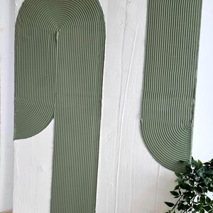 Könnte beinhalten: Abstrakte Wandkunst mit einem strukturierten weißen Hintergrund und zwei grünen, geschwungenen Formen. Die Formen bestehen aus einem strukturierten Material und haben ein erhabenes, gestreiftes Muster.