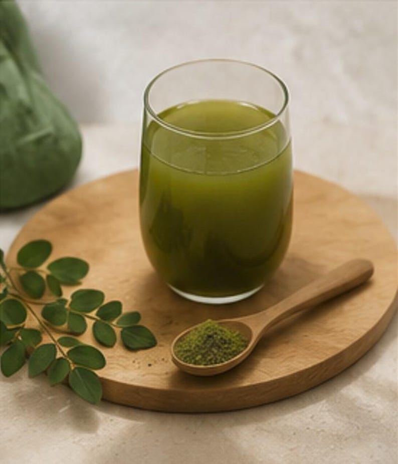 May include: A glass of green liquid sits on a wooden board, next to a wooden spoon filled with green powder. A sprig of green leaves is on the board. The glass is clear and the liquid is opaque.