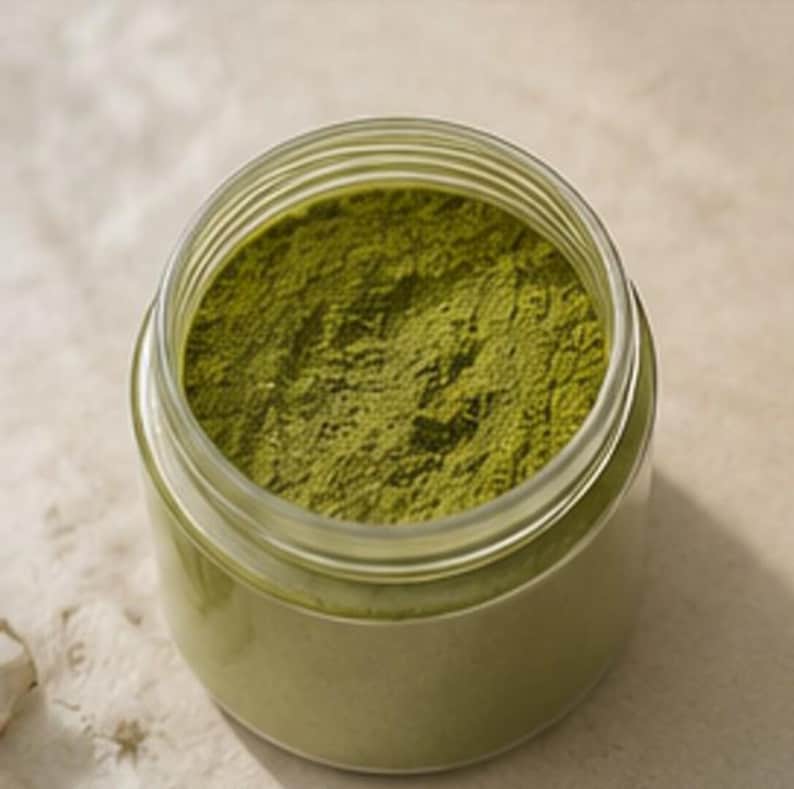 May include: A clear glass jar filled with finely ground, vibrant green powder. The jar has a wide opening and a screw-on lid. The powder appears to be a natural product, possibly a spice or herb, and is a rich, earthy green color.