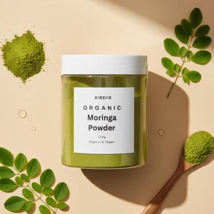 May include: A clear plastic jar of green moringa powder with a white lid and label. The label reads "KIREIIS ORGANIC Moringa Powder 150g Organic & Vegan." Green moringa powder is also on a wooden spoon and scattered on the beige surface. Green moringa leaves are also present.