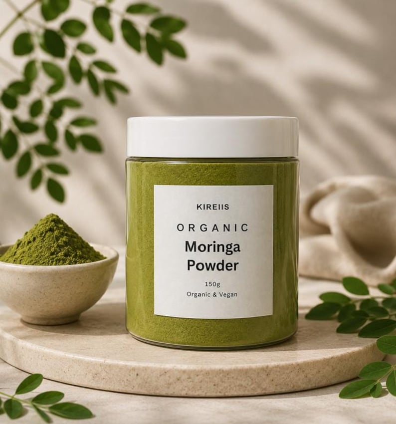 May include: A jar of KIREIIS Organic Moringa Powder, 150g, with a white lid and label. A small bowl of green powder sits to the left. The background includes green leaves and a neutral-toned surface.