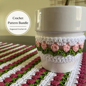 Puede incluir: Una maceta de cerámica blanca con una cubierta de ganchillo con flores rosas y hojas verdes. La maceta está sobre un mantel individual de ganchillo con un patrón a rayas de blanco, rosa y verde.