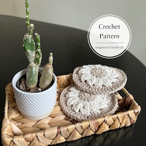 May include: Two crocheted coasters in beige and white with a flower design. The coasters are on a woven tray with a small cactus plant in a white pot.