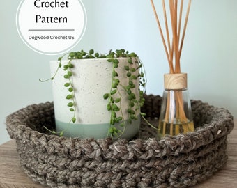 Könnte beinhalten: Ein braunes gehäkeltes Tablett mit einer weißen und grünen Topfpflanze und einer durchsichtigen Glasflasche mit Holzstäbchen darin. Das Tablett steht auf einer Holzoberfläche. Der Text "Crochet Pattern Dogwood Crochet US" befindet sich auf einem weißen Kreis.