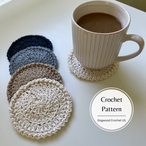 Op de afbeelding: Een set gehaakte onderzetters in neutrale kleuren: crème, beige, grijs en marineblauw. Een witte mok met koffie staat op een onderzetter. De afbeelding bevat de tekst "Crochet Pattern" en "Dogwood Crochet US".