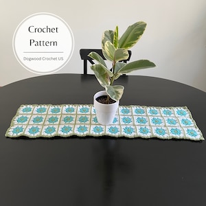 May include: A crocheted table runner with a green and blue floral pattern. The runner is on a black table with a potted plant in the center.