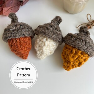 May include: Three crocheted acorns in different colors: orange, white, and yellow. The acorns are made with a brown yarn for the cap and a different color yarn for the body. The text "Crochet Pattern" and "Dogwood Crochet US" is visible in the image.