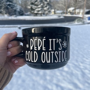 以下が含まれることがあります： 白い文字で「Bébé it's cold outside」と雪の結晶のグラフィックが書かれた黒いセラミックマグカップ。赤と白のマニキュアを塗った手でマグカップを持っています。