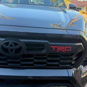 May include: Close-up of a black Toyota truck grille with a black Toyota emblem and red "TRD" lettering. The grille has a honeycomb pattern. The hood of the truck is visible above the grille.