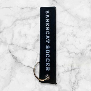 K&ouml;nnte beinhalten: Schwarzer Schl&uuml;sselanh&auml;nger mit dem wei&szlig;en Aufdruck "SABERCAT SOCCER".