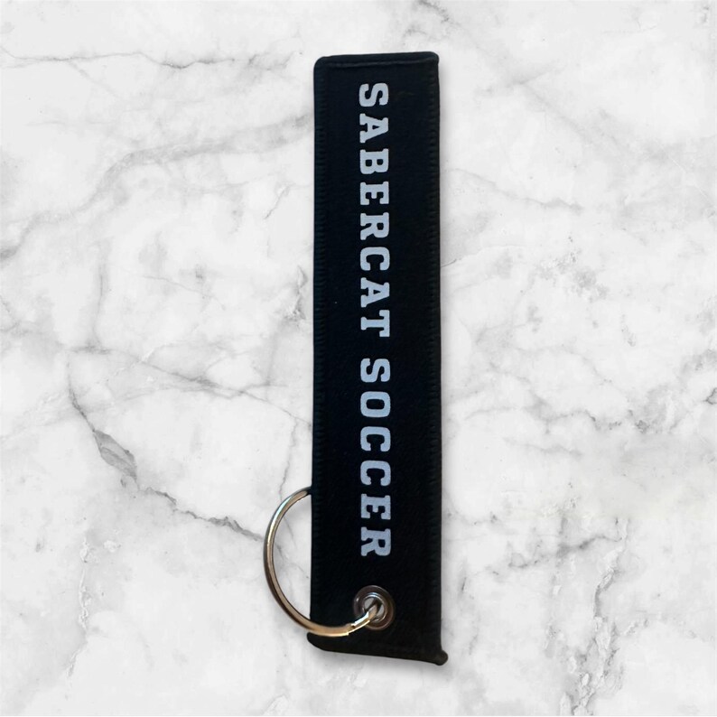 K&ouml;nnte beinhalten: Schwarzer Schl&uuml;sselanh&auml;nger mit dem wei&szlig;en Aufdruck "SABERCAT SOCCER".