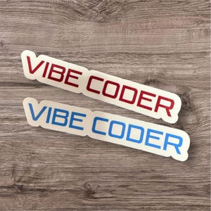 May include: Two stickers with the text "VIBE CODER" in red and blue, respectively. The stickers are rectangular with rounded corners and a white border. The background is a wooden surface.