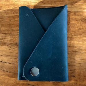 May include: A navy blue leather card holder with a unique folded design. The wallet is secured with a silver snap closure. The card holder is displayed on a wooden surface.