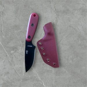 Puede incluir: Un cuchillo Izula negro con mango y funda burdeos. El cuchillo tiene un pequeño logotipo de hormiga en la hoja.