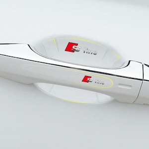 May include: A close-up of a white car door handle with a chrome finish. The handle has a protective film with the red and silver "S line" logo. The background is a blurred white.